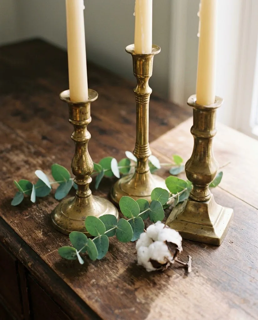 Styled dinner table centerpiece with five taper candle holders in mixed heights on a natural linen runner, loose eucalyptus and dried botanicals between holders, warm candlelight glow, small apartment dining room
